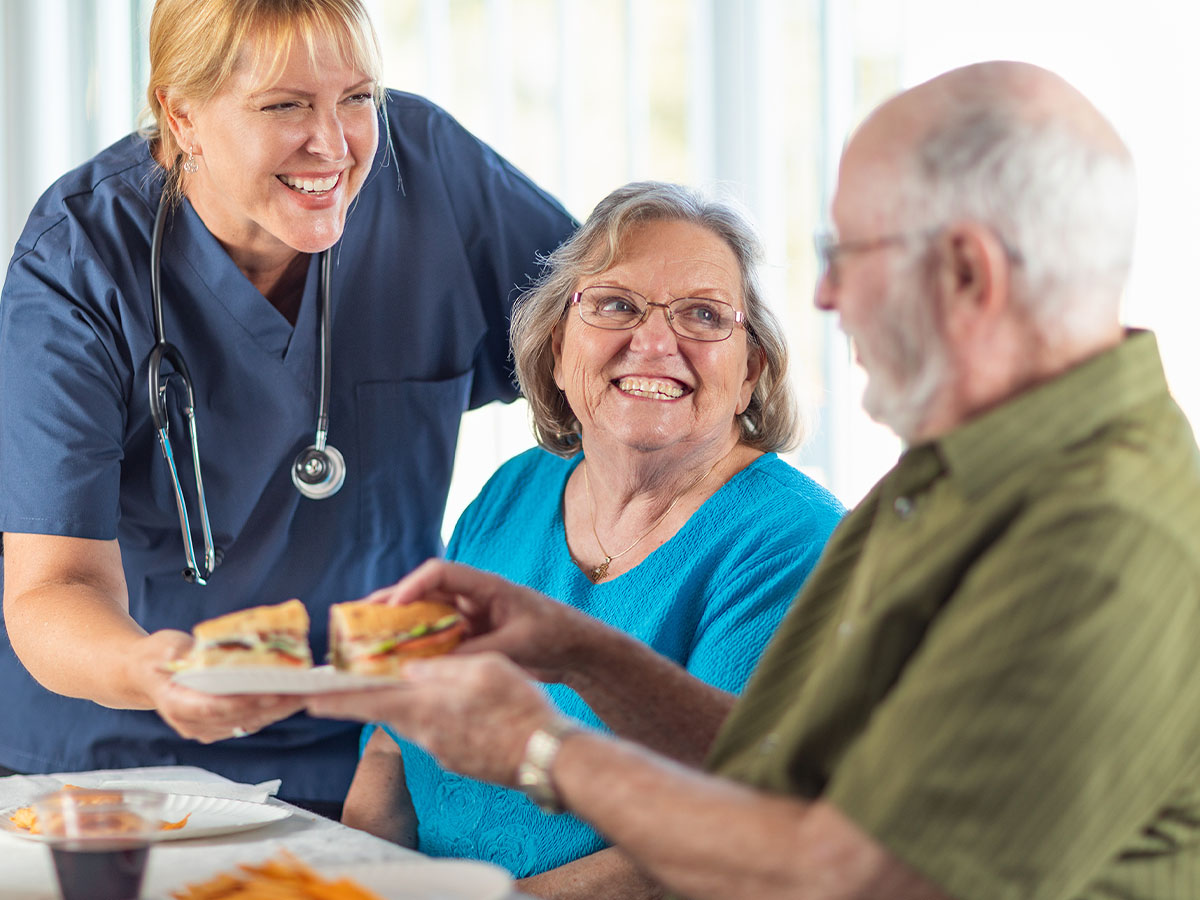 Caregiver handing an elderly man a sandwich while his wife smiles at him