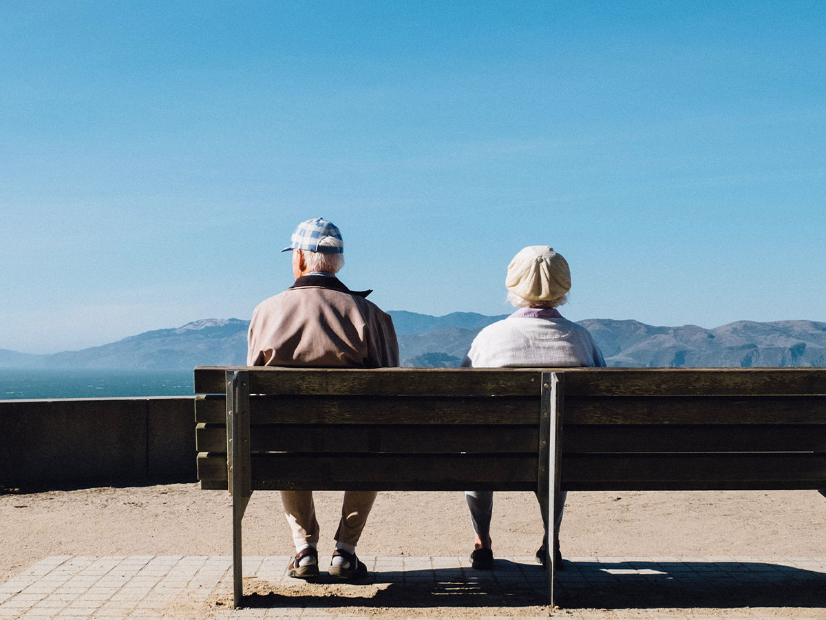 Two elderly people sitting on a bench looking at an ocean and mountainous view