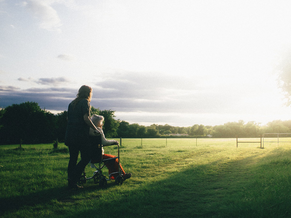 A caregiver pushes an elderly person in a wheelchair through a field